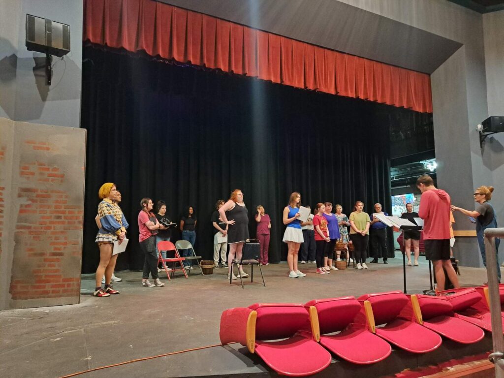 The cast of Cinderella standing in a semi-circle onstage around the choreographer during rehearsal.