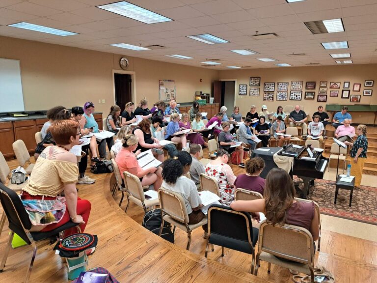 A large group of cast members seated in a rehearsal room, reading from scripts