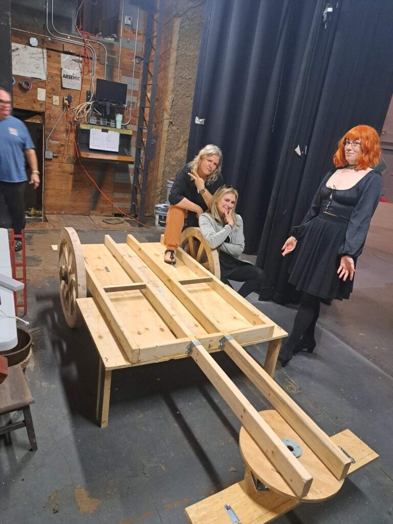 Cast members posing with partially built wooden carriage backstage at Cinderella rehearsals.