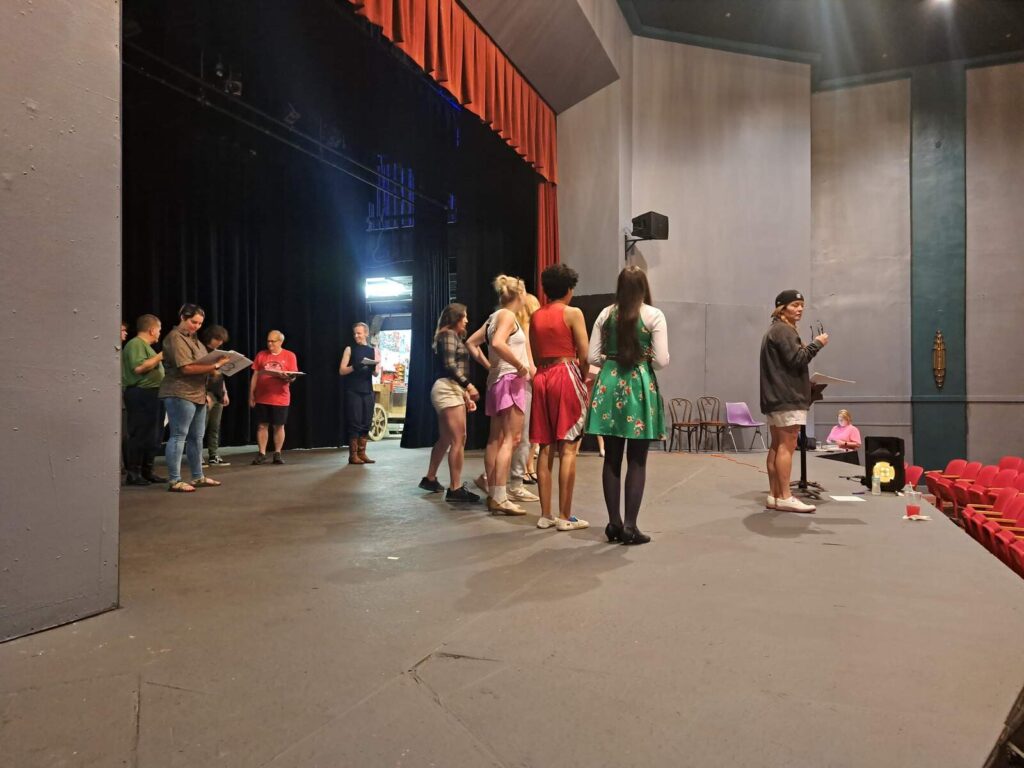 A group of cast members stands on stage while a director gives notes with a music stand in front of her.