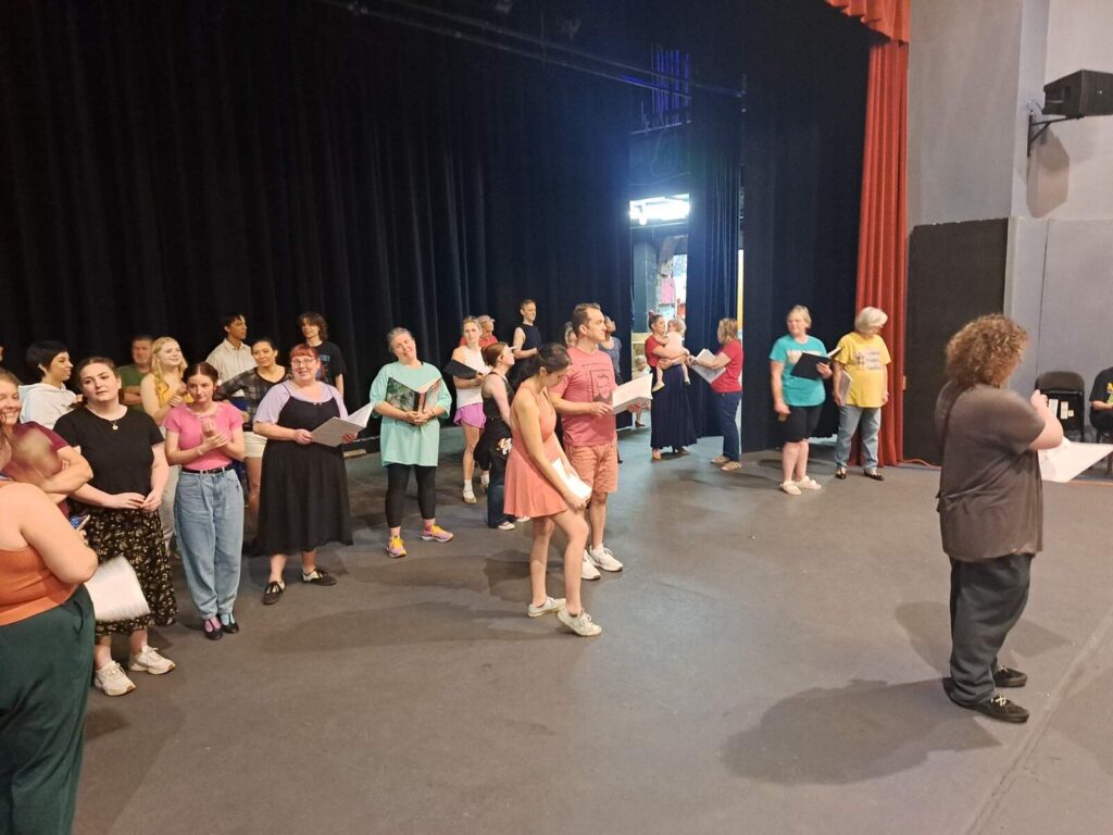 A large group of performers stands in a semicircle onstage while a director gives instructions.