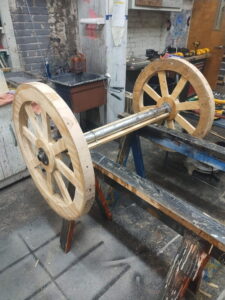 Wooden carriage wheels with a central axle in a workshop, part of the set construction for Tupelo Community Theatre's Production of Rodgers + Hammerstein’s Cinderella.