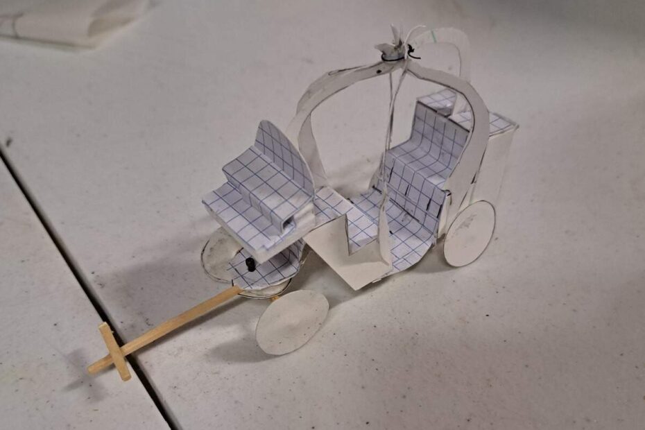 Small hand‑cut paper model of ornate carriage with graph‑paper seats on white table.
