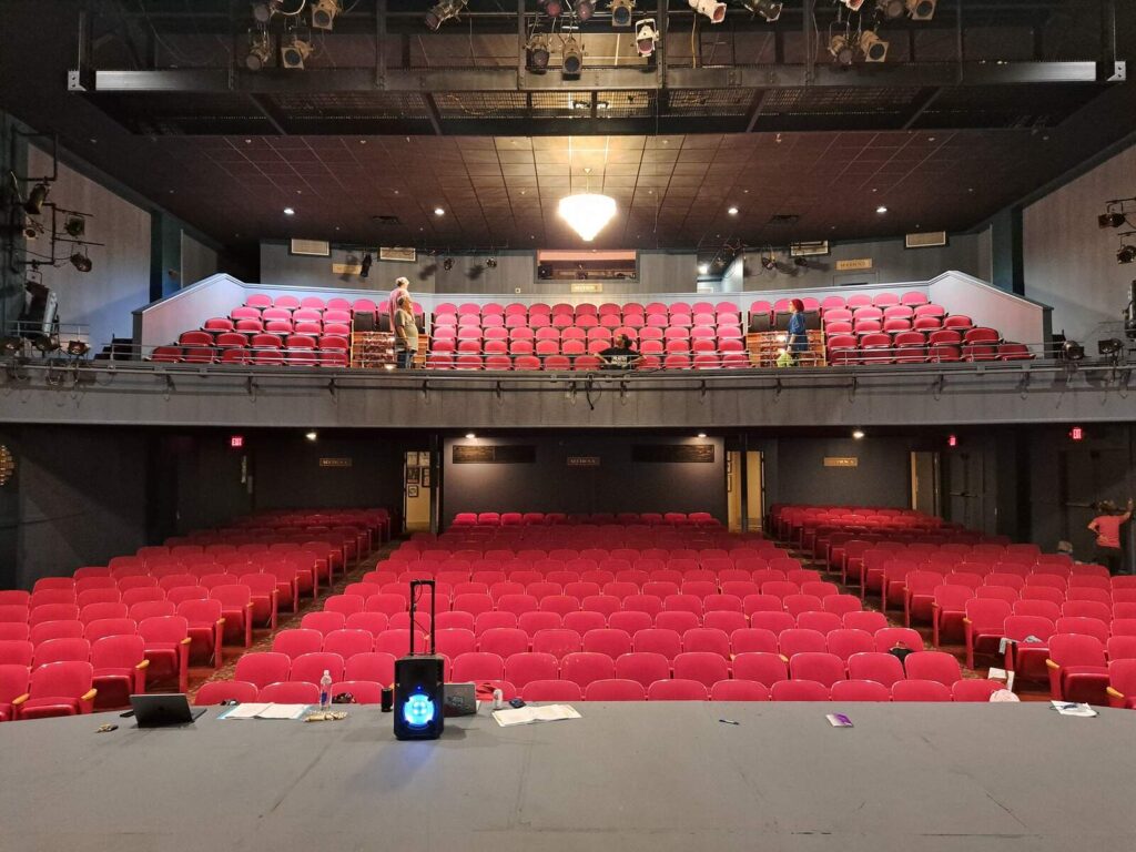View from stage over rows of crimson chairs toward balcony, technicians at work.