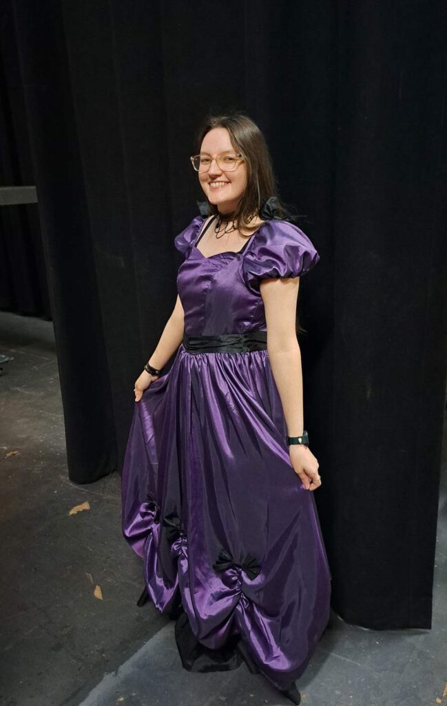 A smiling cast member models a lustrous purple ball gown with puffed sleeves backstage at the Lyric Theatre