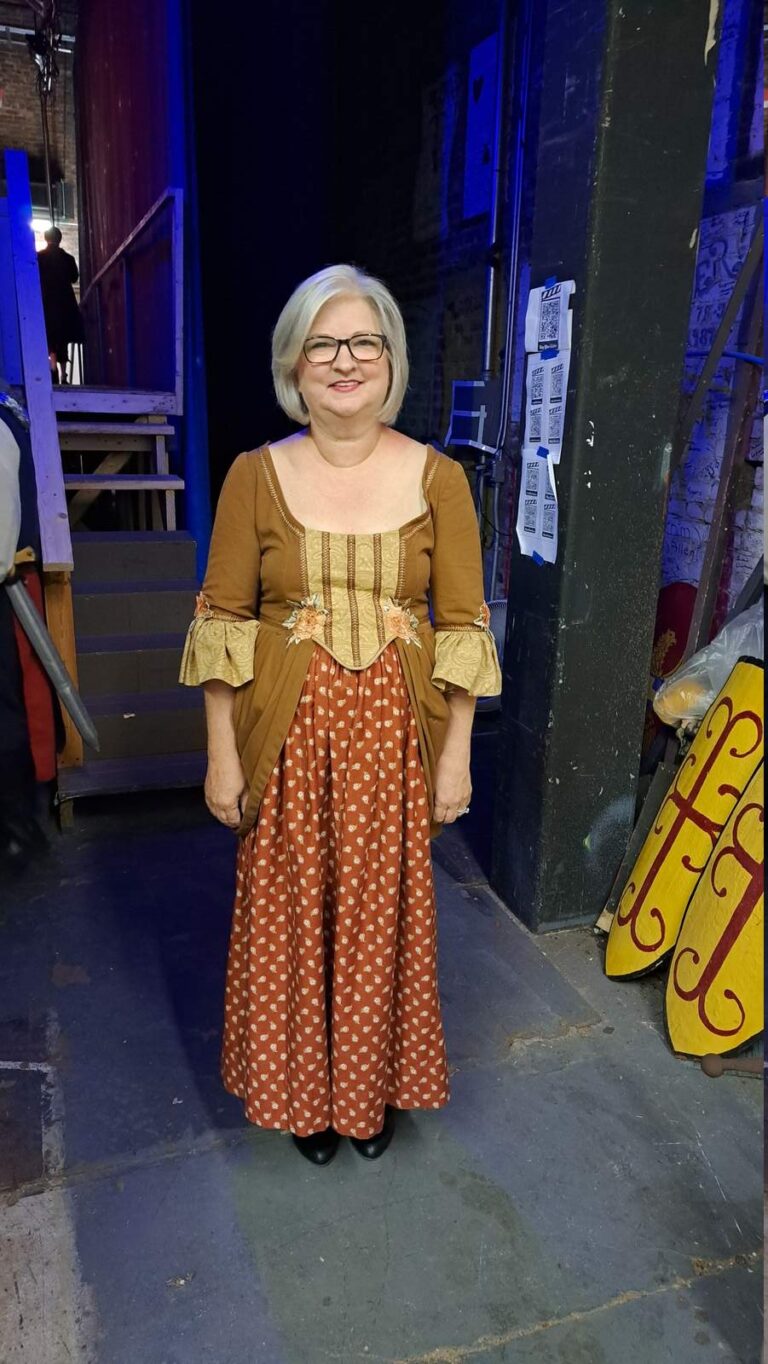 Woman in brown bodice and patterned skirt stands under blue work lights
