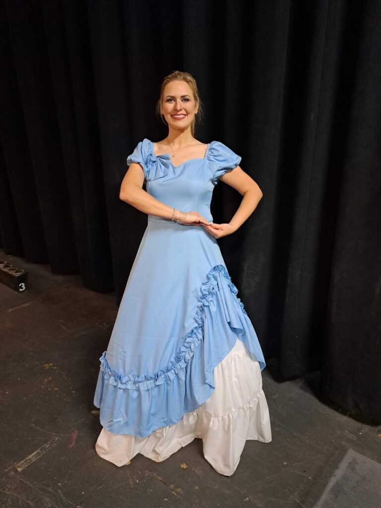 Erin in a blue dress performs a ballroom pose on rehearsal stage.