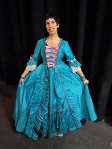 Marina in elaborate teal silk dress with tiered ruffles and pink accents poses before black curtains