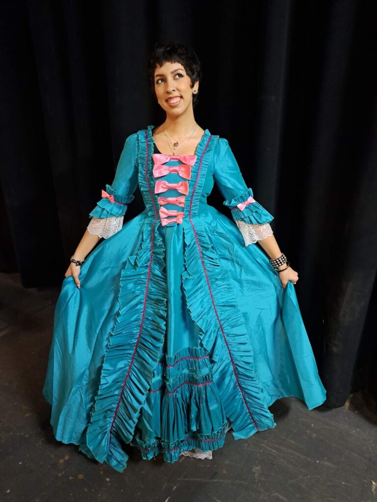 Marina in elaborate teal silk dress with tiered ruffles and pink accents poses before black curtains