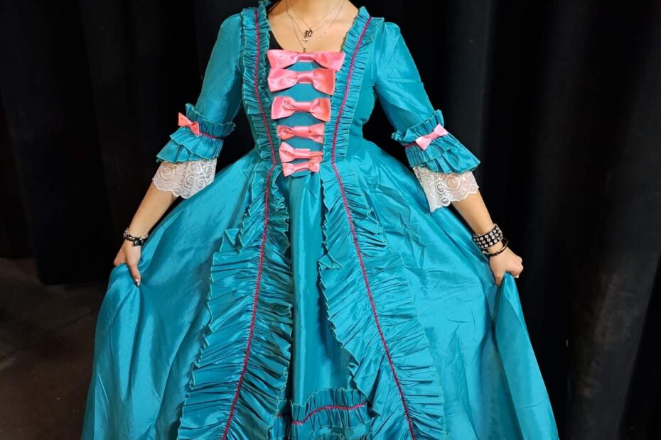 Marina in elaborate teal silk dress with tiered ruffles and pink accents poses before black curtains