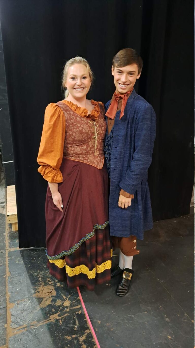 Female actor in ruffled bodice and male actor in blue frock coat pose side-by-side on rehearsal floor.