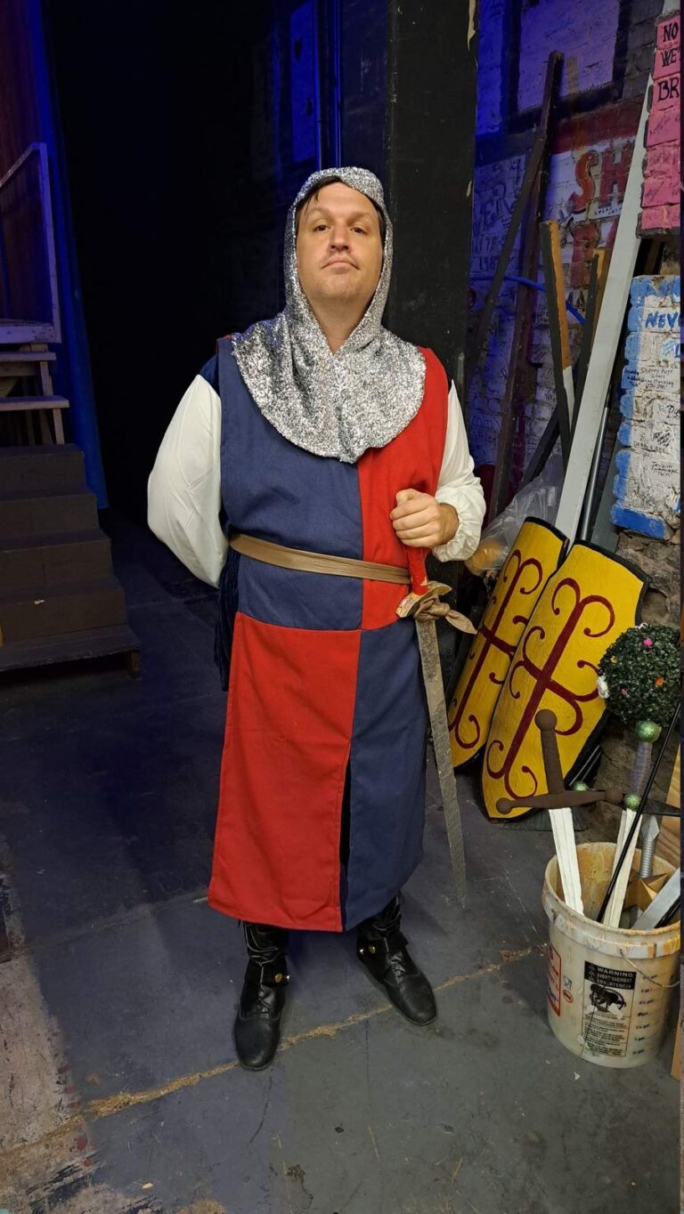 Actor in red-and-navy surcoat holds sword backstage