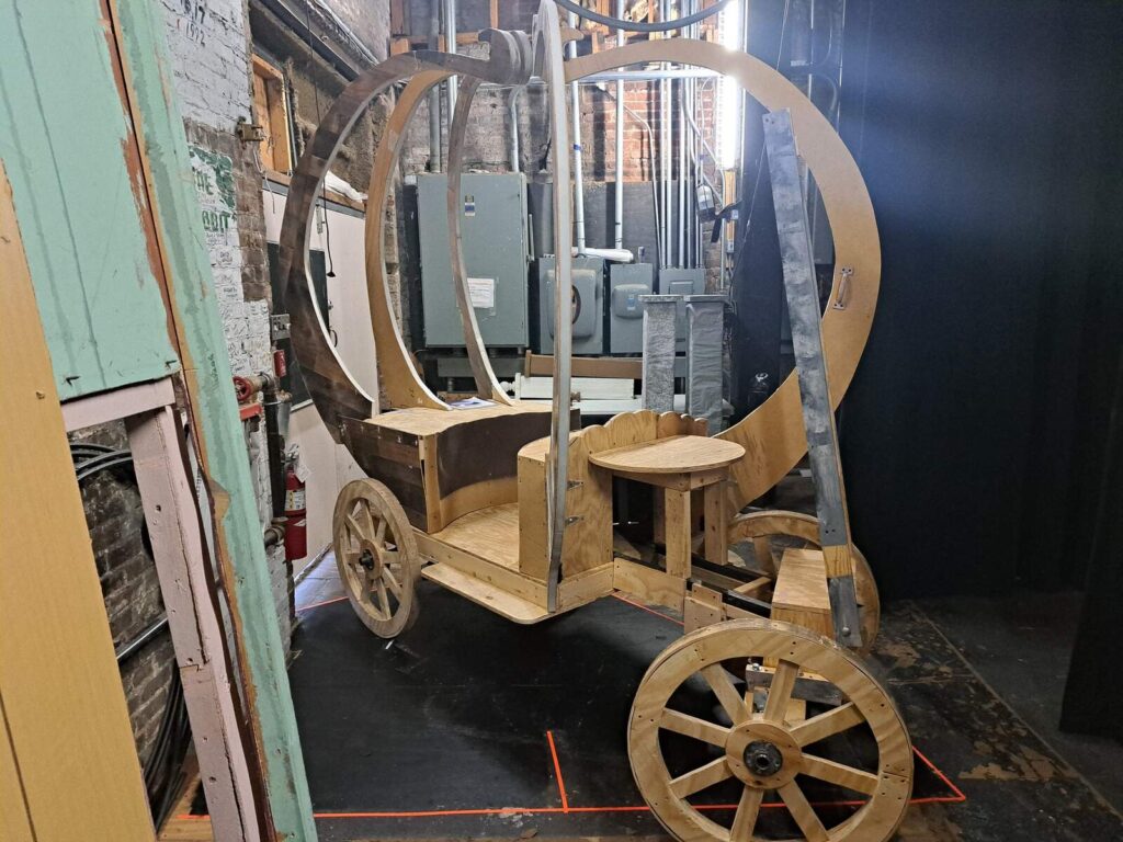 Unpainted wooden carriage frame shaped like a pumpkin rests on sturdy wheels