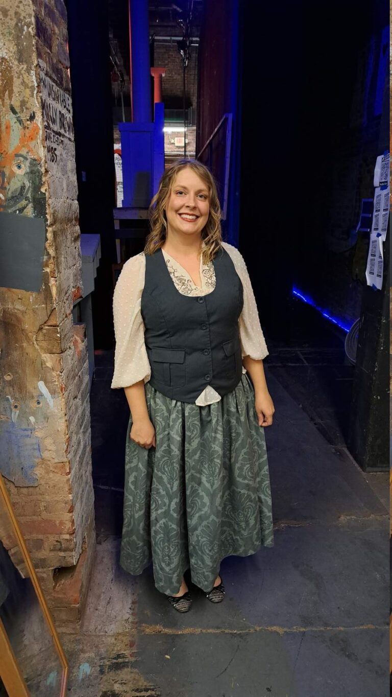 Cast member in muted village dress poses by brick column