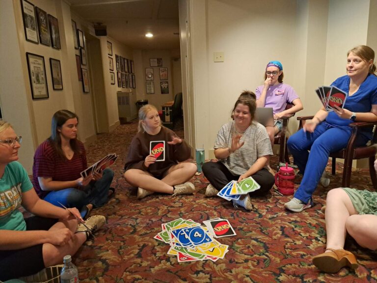 Circle of performers seated on carpet, holding oversized UNO cards during a break.