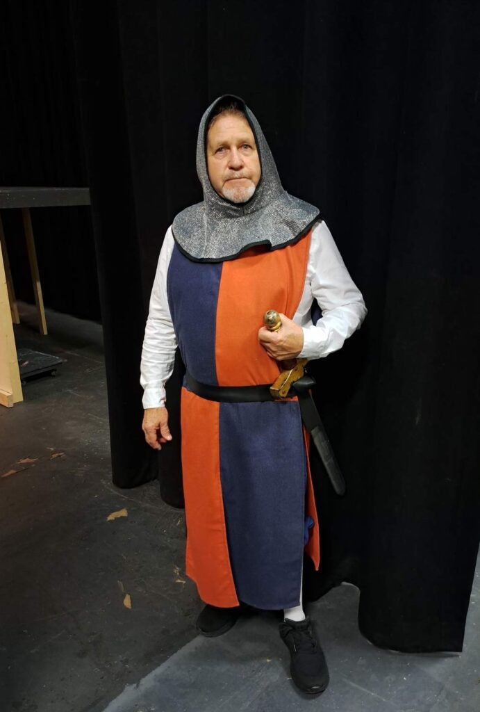 Cast member wears a split blue-and-rust tunic, chain-hood, and sword