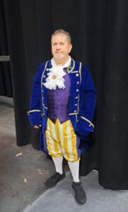 Cast member in cobalt velvet coat, ruffled cravat, and striped yellow breeches stands center stage.