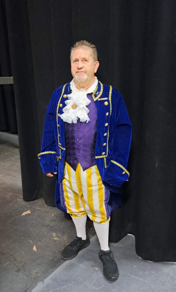 Cast member in cobalt velvet coat, ruffled cravat, and striped yellow breeches stands center stage.