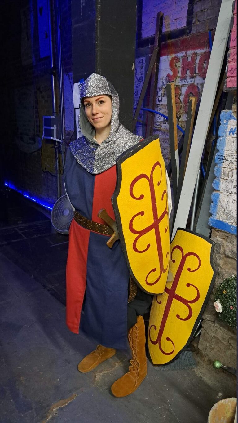 Cast member in silver coif and blue-and-red tunic poses with a bright yellow shield behind the wings
