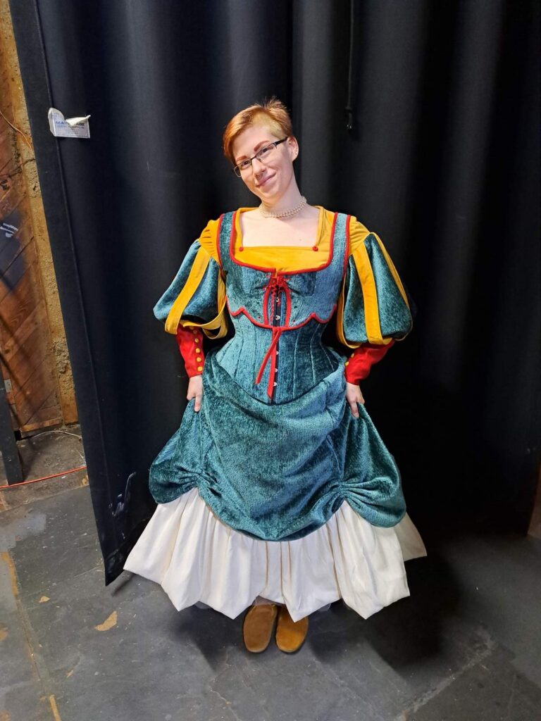 Cast member displays a teal velvet dress with golden-yellow sleeves and red lacing, holding the skirt sides