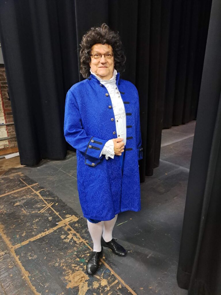 Man in 18th-century blue coat stands on stage