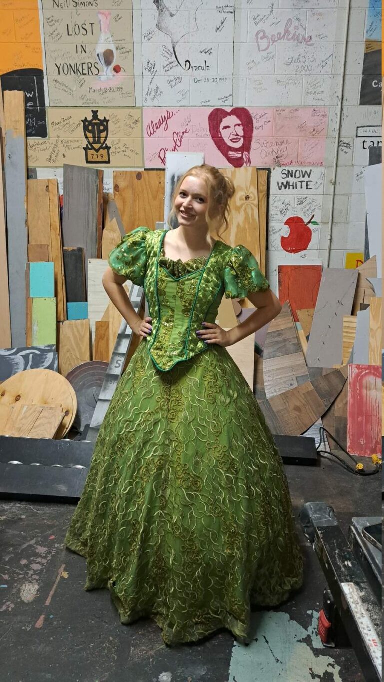 Performer wearing ornate green gown with puffed sleeves and gold embroidery stands hands-on-hips backstage