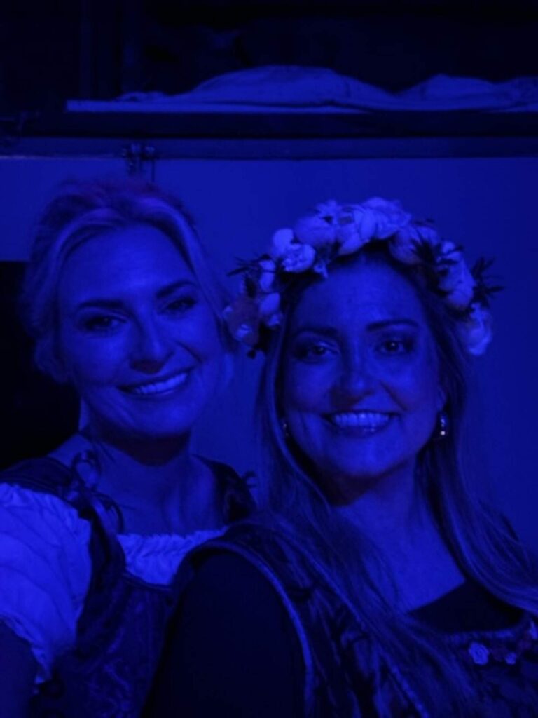 Two female cast members in peasant dresses smile under deep blue work-light just offstage