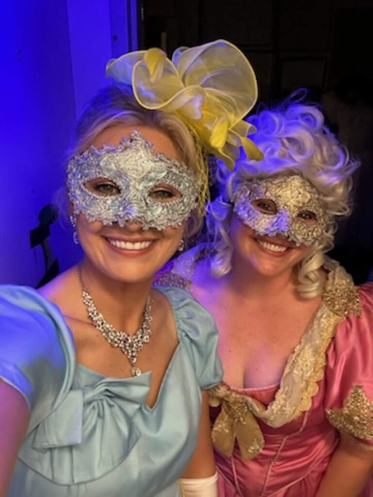 Two women in ornate gowns and lace masks pose in soft backstage light