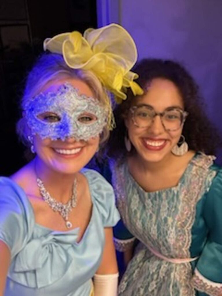 Erin in blue gown and filigree mask poses beside Kennedy in teal brocade dress
