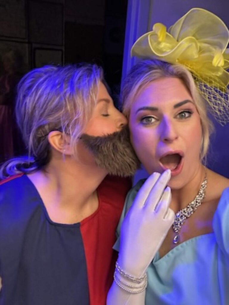 Actress in faux beard kisses surprised masked lady in blue ball gown backstage.
