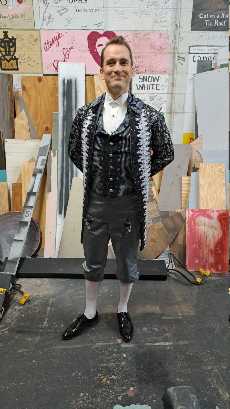 Performer in charcoal knee-breeches and embroidered jacket stands hands-behind-back