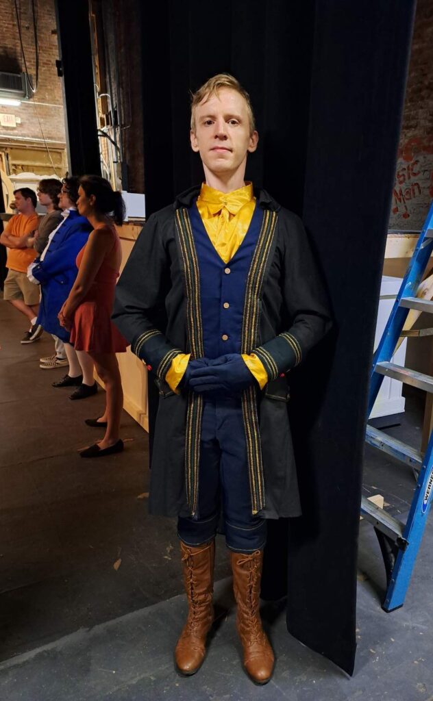 Cast member stands tall in black coat with gilded trim and bright yellow undershirt beside the wing