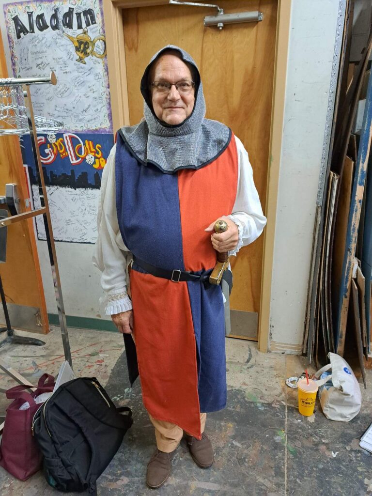 Bobby Geno in red-and-blue tabard with chainmail-style hood holding sword hilt