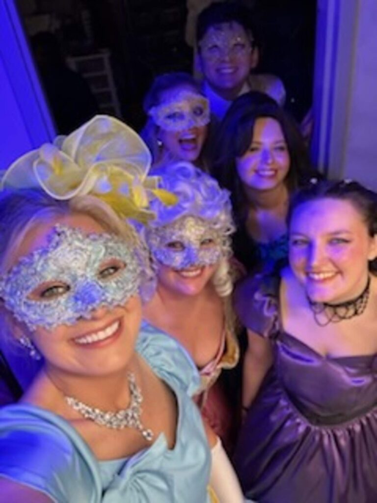 Cluster of smiling performers in courtly attire framed by backstage doorway.