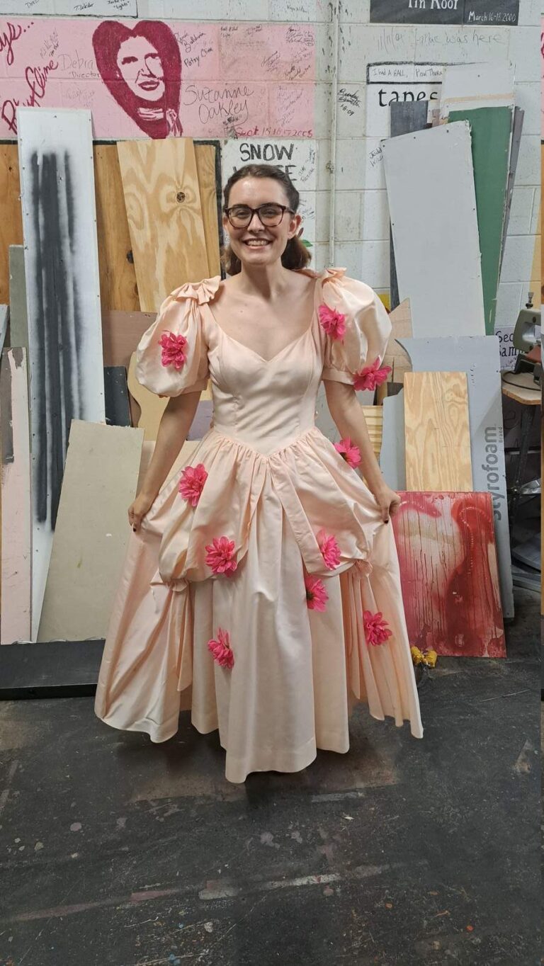 Actor in full-length peach gown decorated with silk blossoms backstage