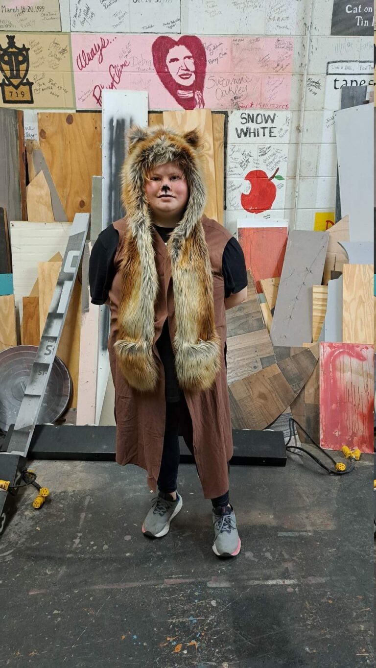Performer wears brown vest with long faux-fur hood and painted nose