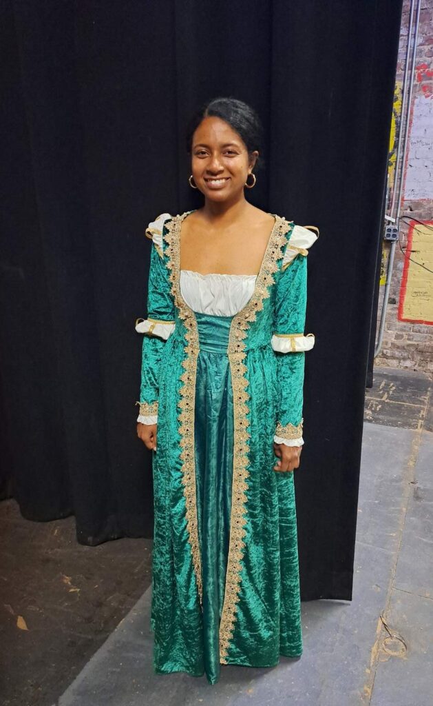 Cast member smiles in a long dark-green velvet gown with lace trim and gathered sleeves