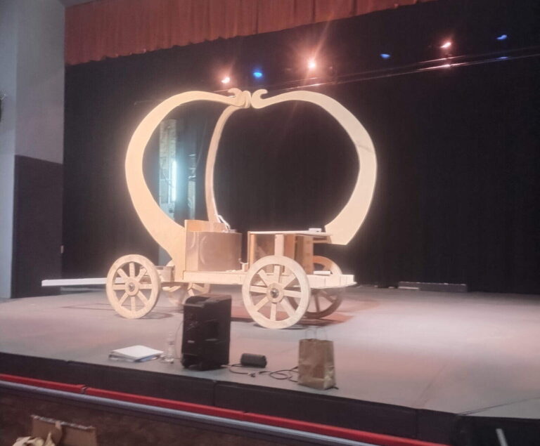 Wide shot of light-wood carriage base with large wheels on empty theater stage, red seats in foreground.