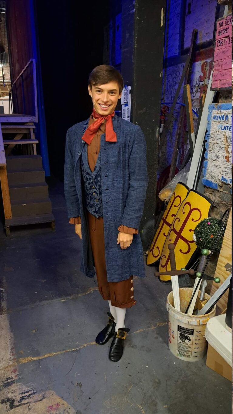 Performer in long indigo coat, brown breeches, and red neckerchief smiles