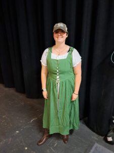 Actor in green medieval-style dress, white peasant blouse, brown boots, and gray ball cap on rehearsal stage