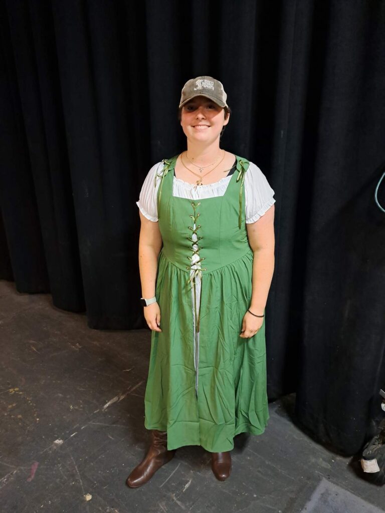 Actor in green medieval-style dress, white peasant blouse, brown boots, and gray ball cap on rehearsal stage