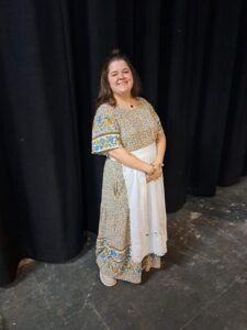 Actor in ankle-length floral dress with white apron, smiling broadly on stage.