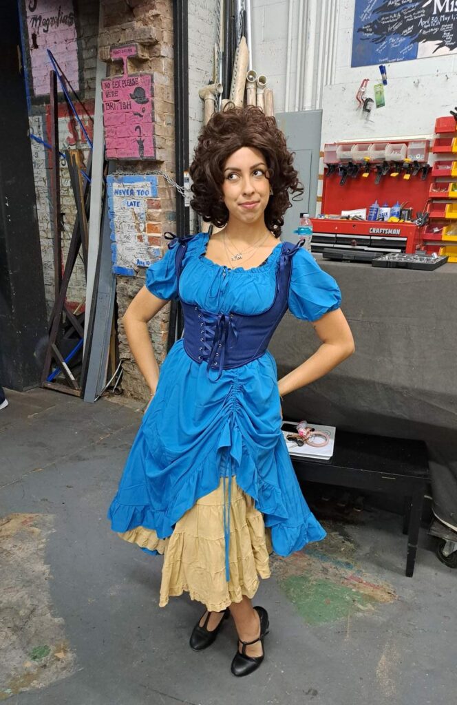 Actor standing backstage in deep blue bodice and puff-sleeve blue dress.
