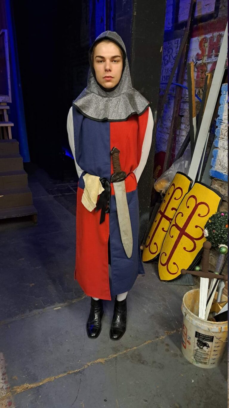 Stoic guard in blue-and-scarlet tunic, black boots and chain hood stands on dim stage wing
