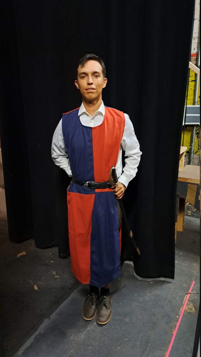 Actor in red-and-navy tabard over white shirt, leather belt and prop sword stands before black curtain